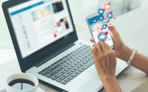 Person holding cell phone with social media icons on the screen, with open laptop in the background.