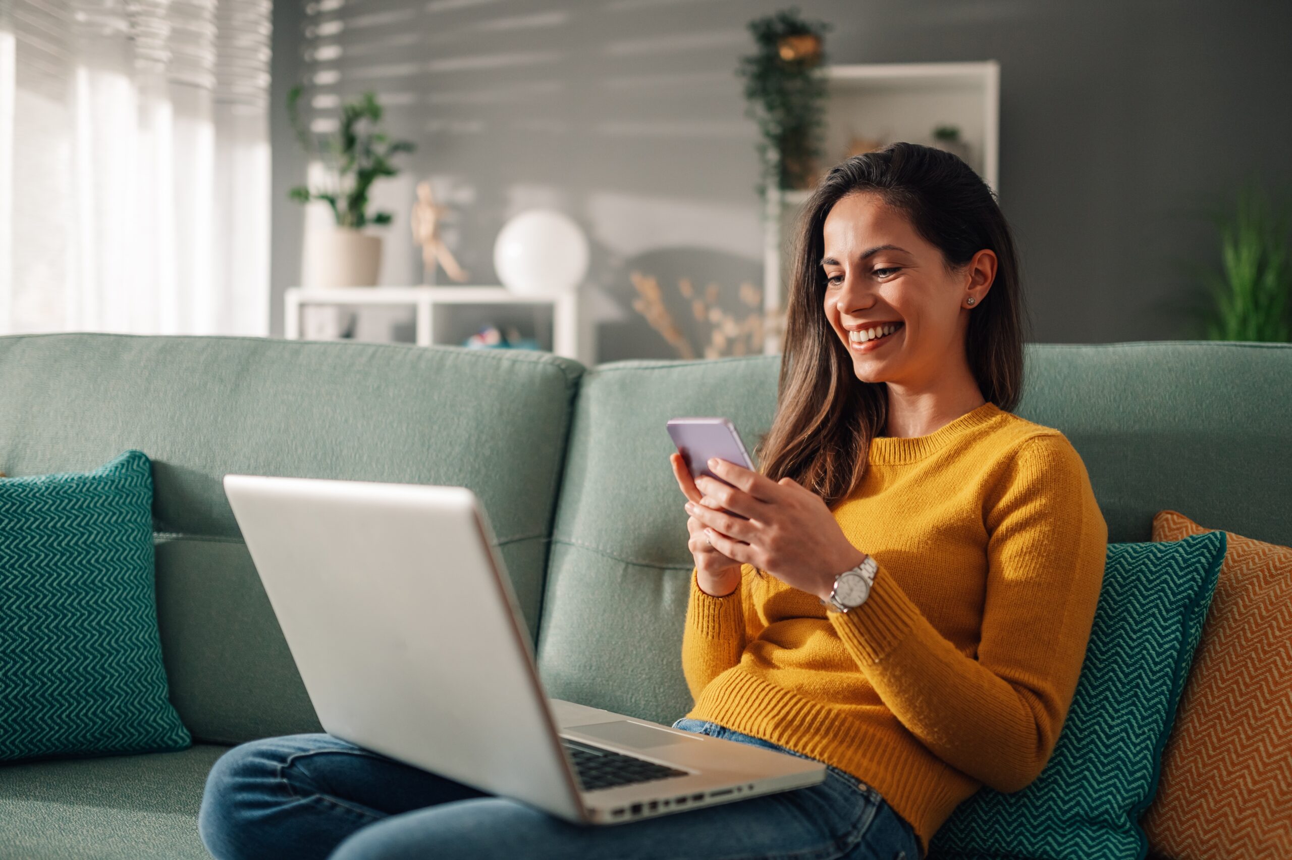 Woman sitting on a couch with an open laptop on her lap, looking at a cell phone.