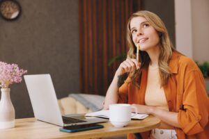 Woman copywriter at table with laptop, tablet, and mug, looking off as if she is thinking